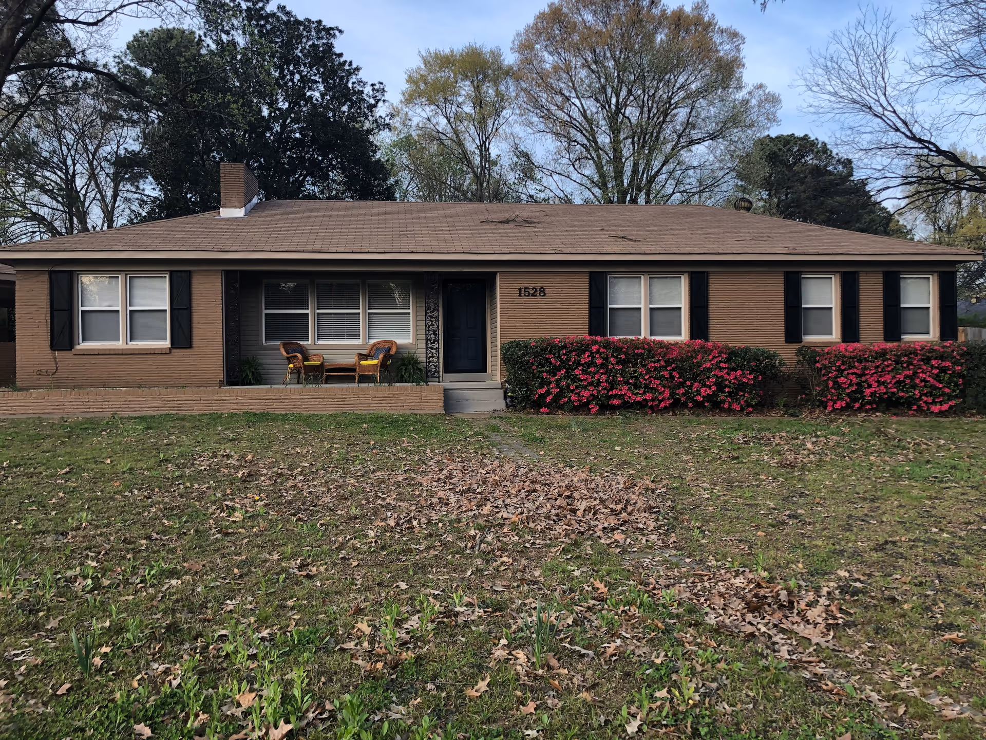 Single-story brick house with front porch chairs and pink flowering shrubs.