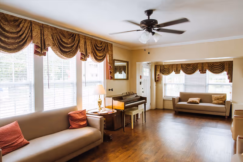 Sunlit communal living room with sofas, a piano, large windows with draped valances, a side table lamp and ceiling fan.