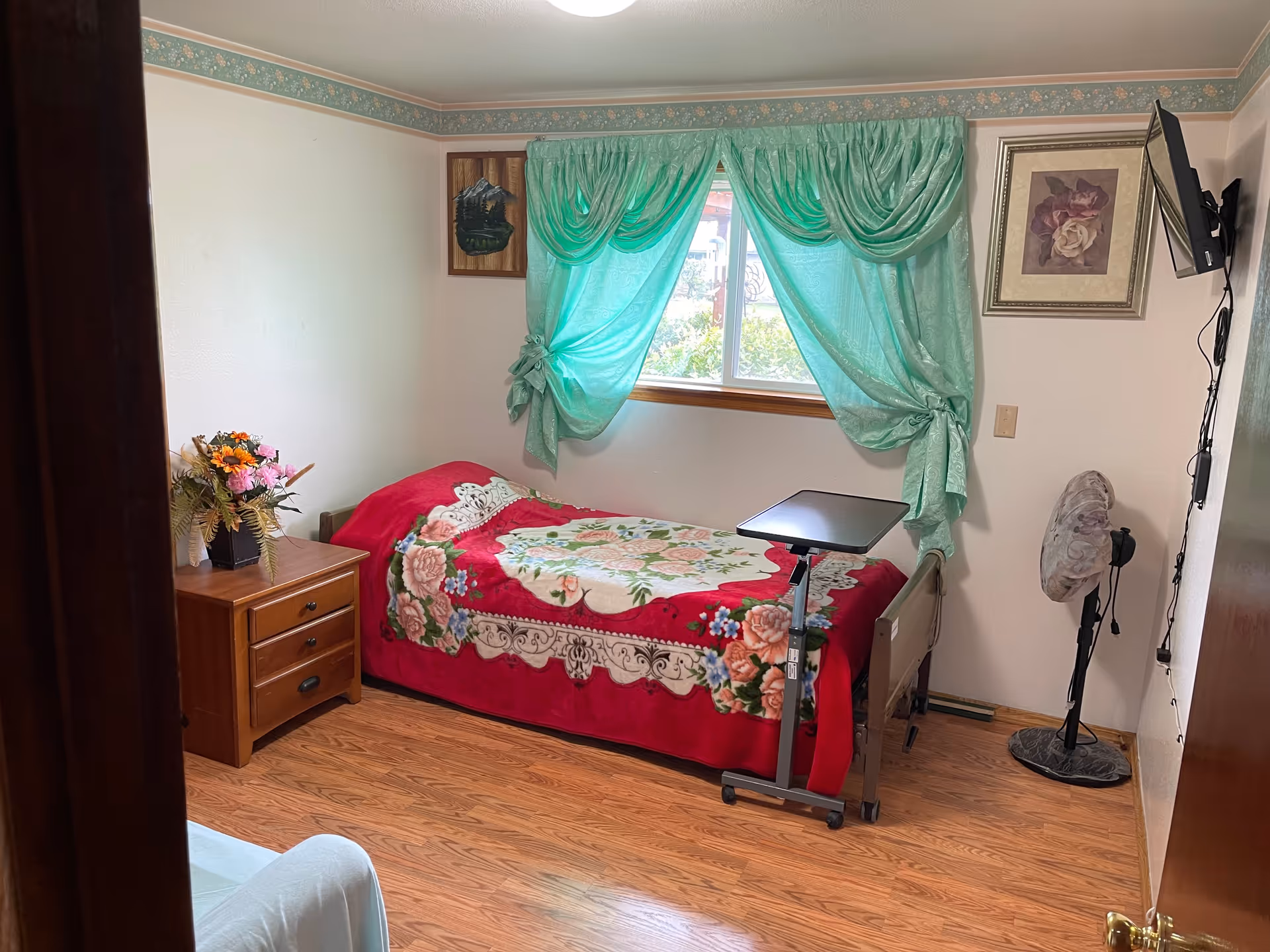 A small bedroom with a floral bedspread, green curtains, a nightstand, and a TV.
