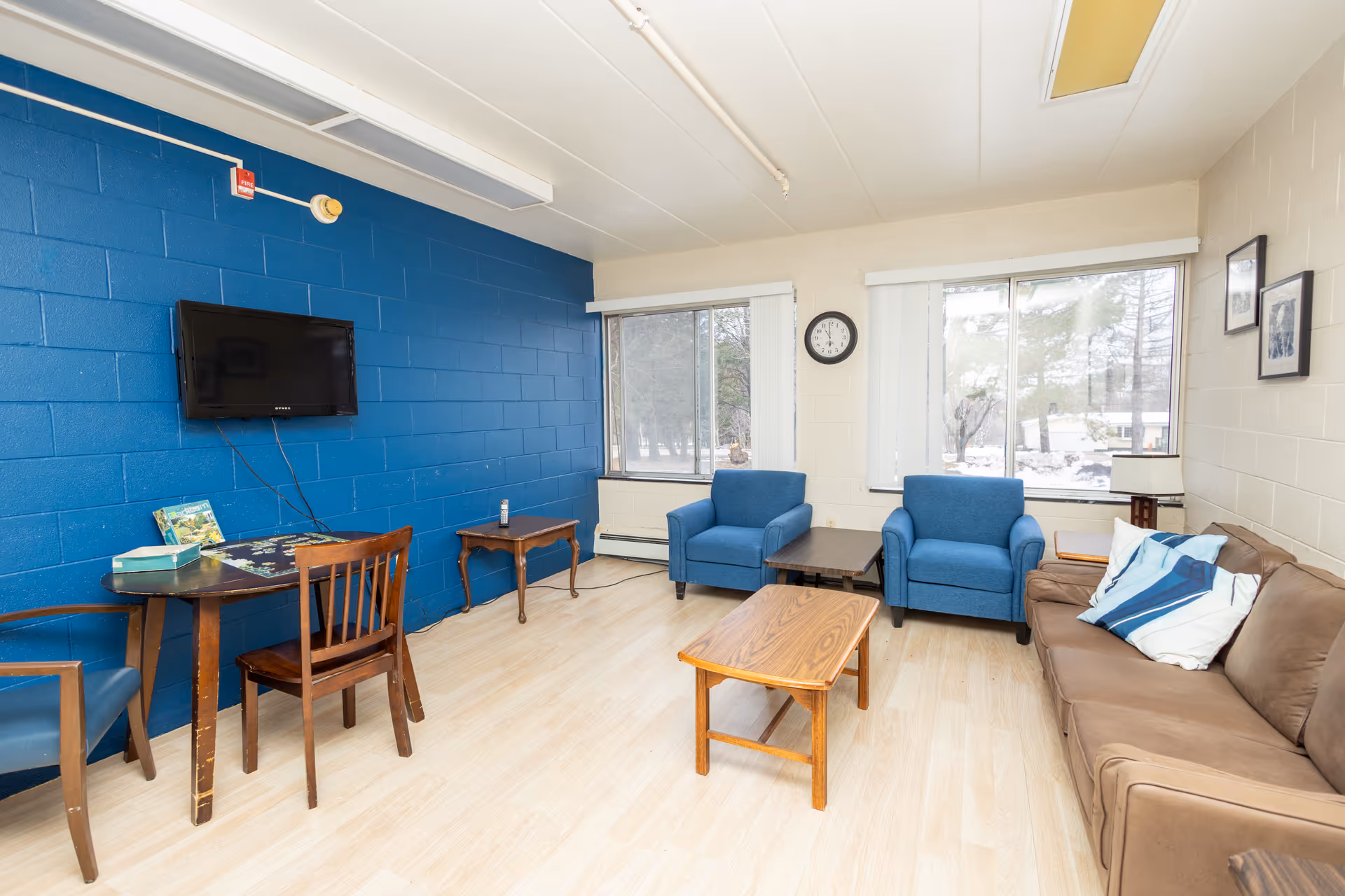 A bright living room with light wood flooring, a blue accent wall with a mounted flat-screen TV, two blue armchairs, a brown sofa with pillows, a wooden coffee table, a small side table, and a round table with chairs. Large windows let in natural light and show a snowy outdoor scene.