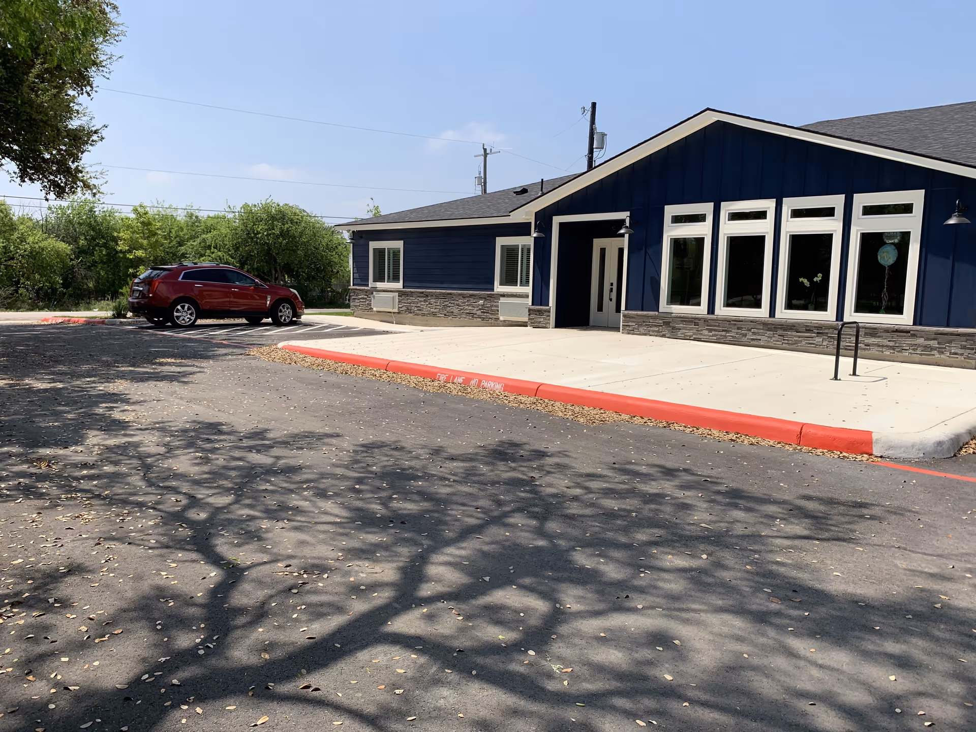 Front exterior of a single-story blue building with large windows, a concrete entryway and a nearby parked red SUV in the parking lot.