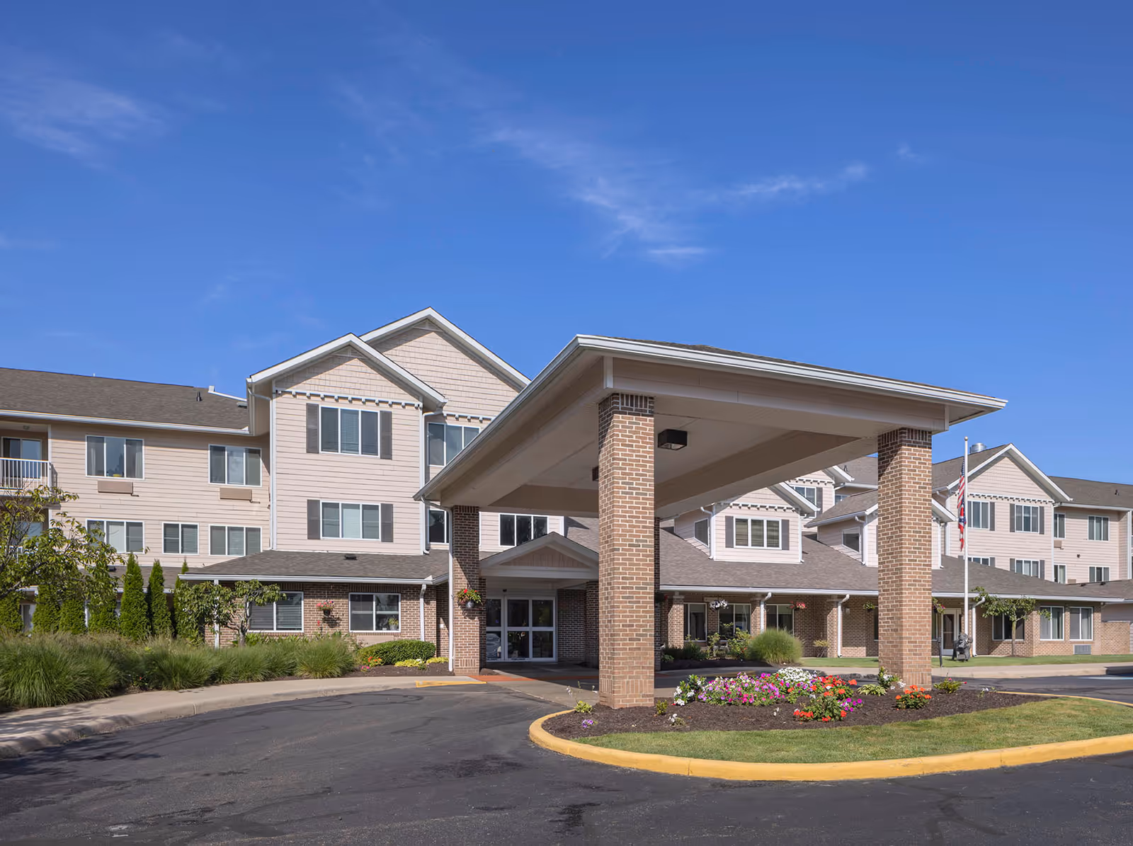 Exterior view of Holiday Copley Place, a multi-story senior living facility with beige siding and brick accents. The entrance features a covered drop-off area supported by brick columns, surrounded by landscaped flower beds and greenery under a clear blue sky.