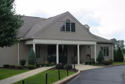 Beige single-story building with a covered columned entrance, walkway, shrubs, and a paved driveway.