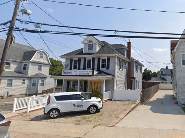 Two-story gray house with Senior Helpers sign, a white company van, and street view.