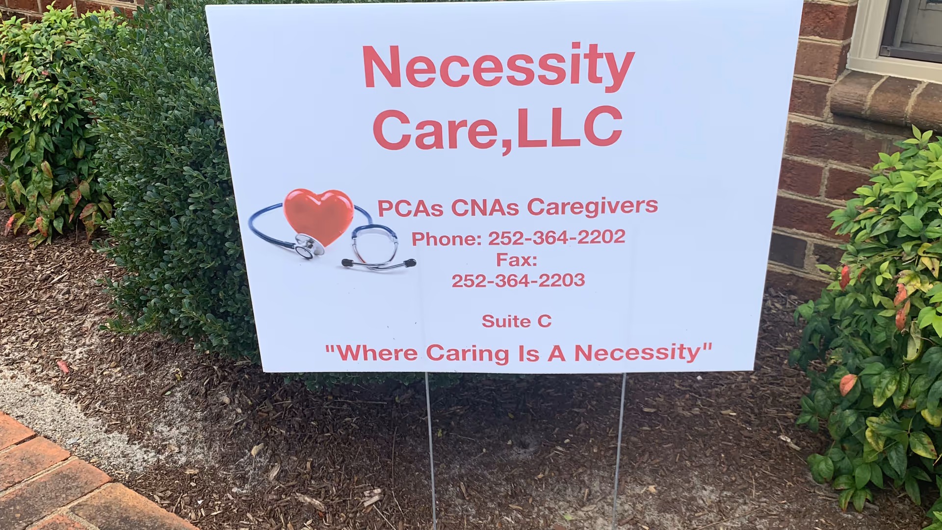 A white sign for Necessity Care, LLC with a heart and stethoscope outside.