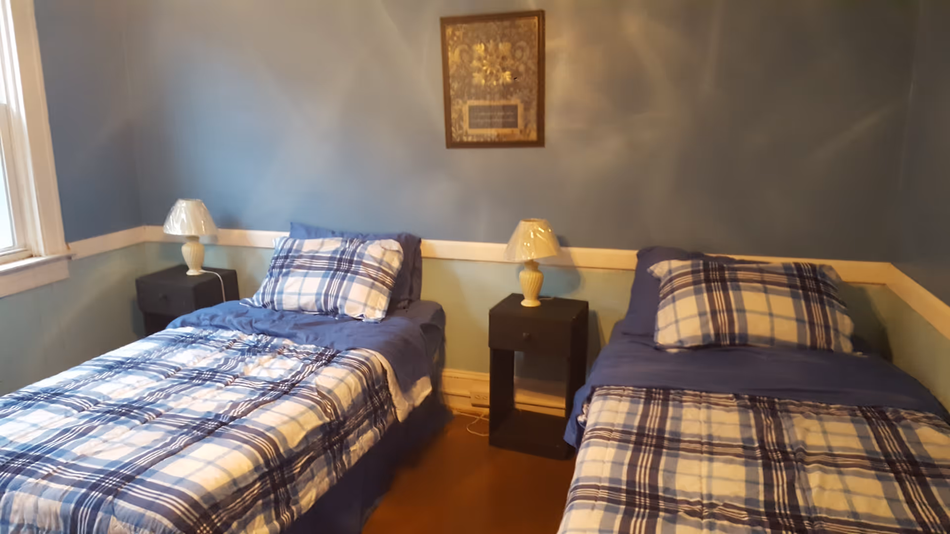 A bedroom with two single beds placed side by side, each with blue and white plaid bedding and pillows. Between the beds is a small black nightstand with a cream-colored lamp on top. The walls are painted blue with a white trim, and a framed picture hangs above the nightstand. There is a window on the left side of the room.