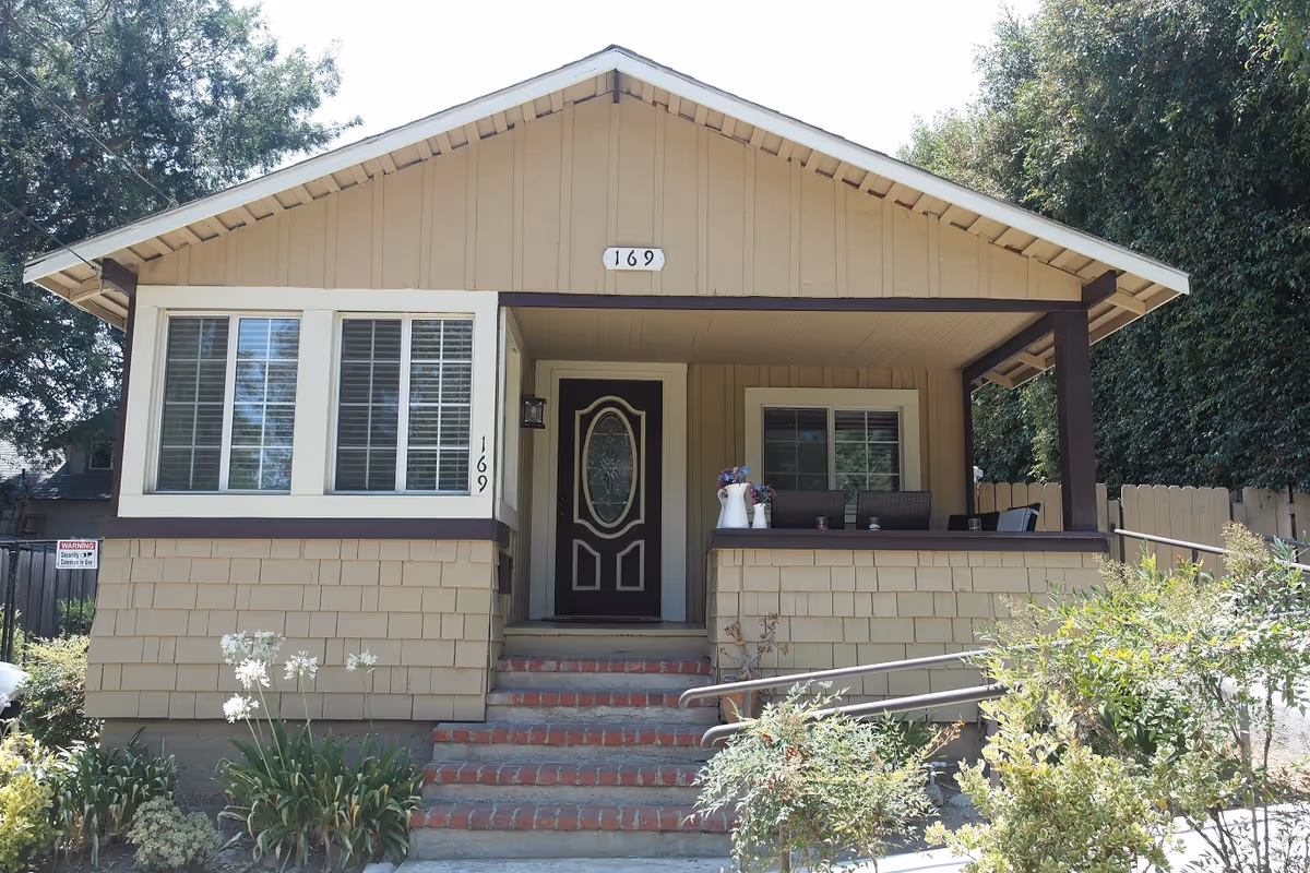 Front view of a beige single-story cottage with a covered porch, steps up to a black front door marked 169, and plants in the yard.