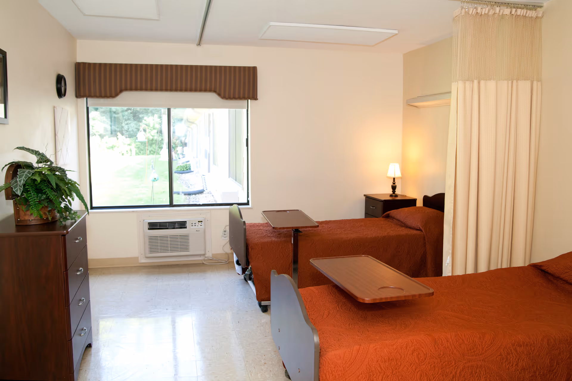 A two-bed nursing room with red bedspreads, over-bed tables, a window with an air conditioner, dresser and a privacy curtain.