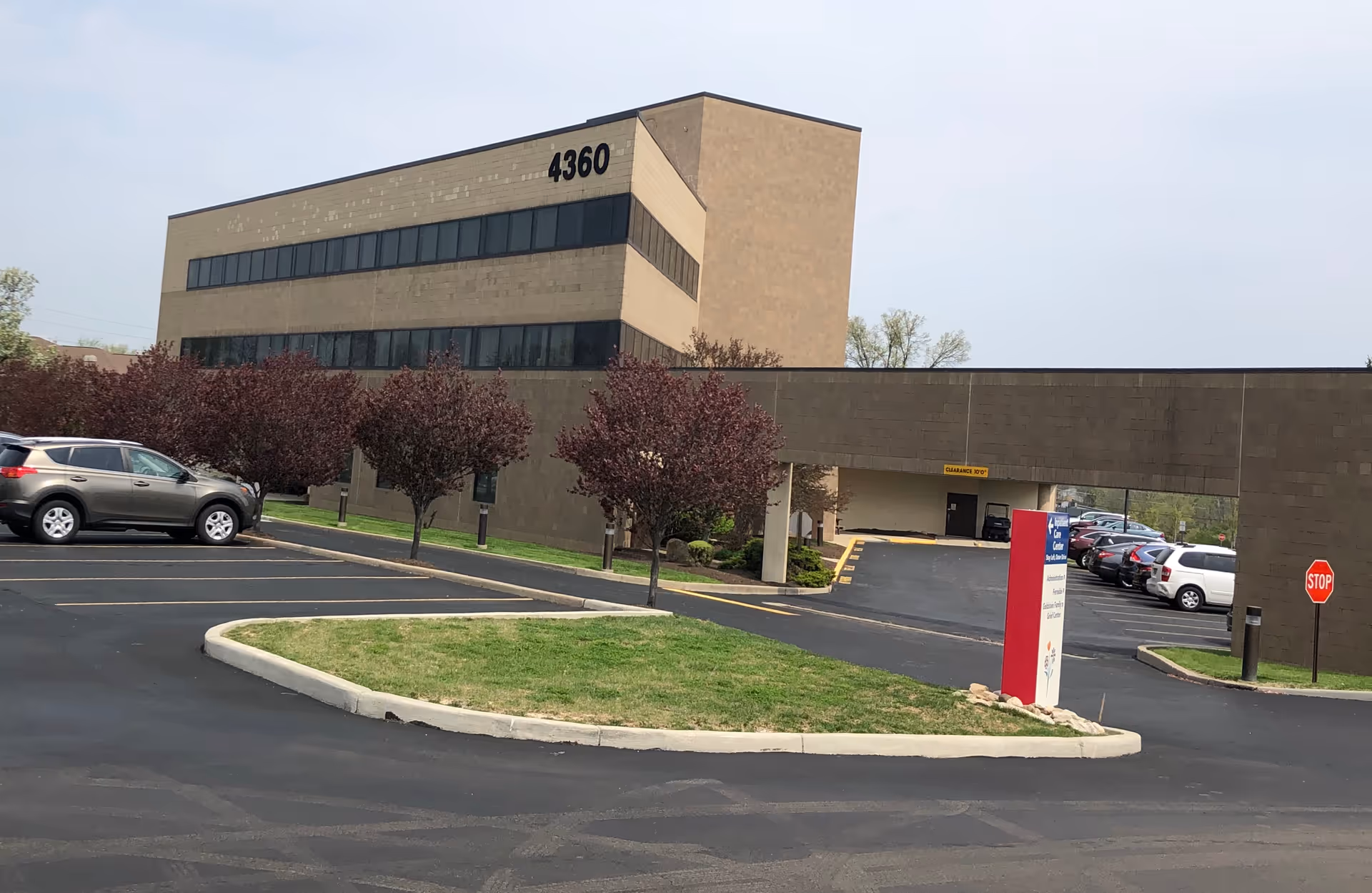 Large beige office building with a parking lot, trees, and a red information sign.