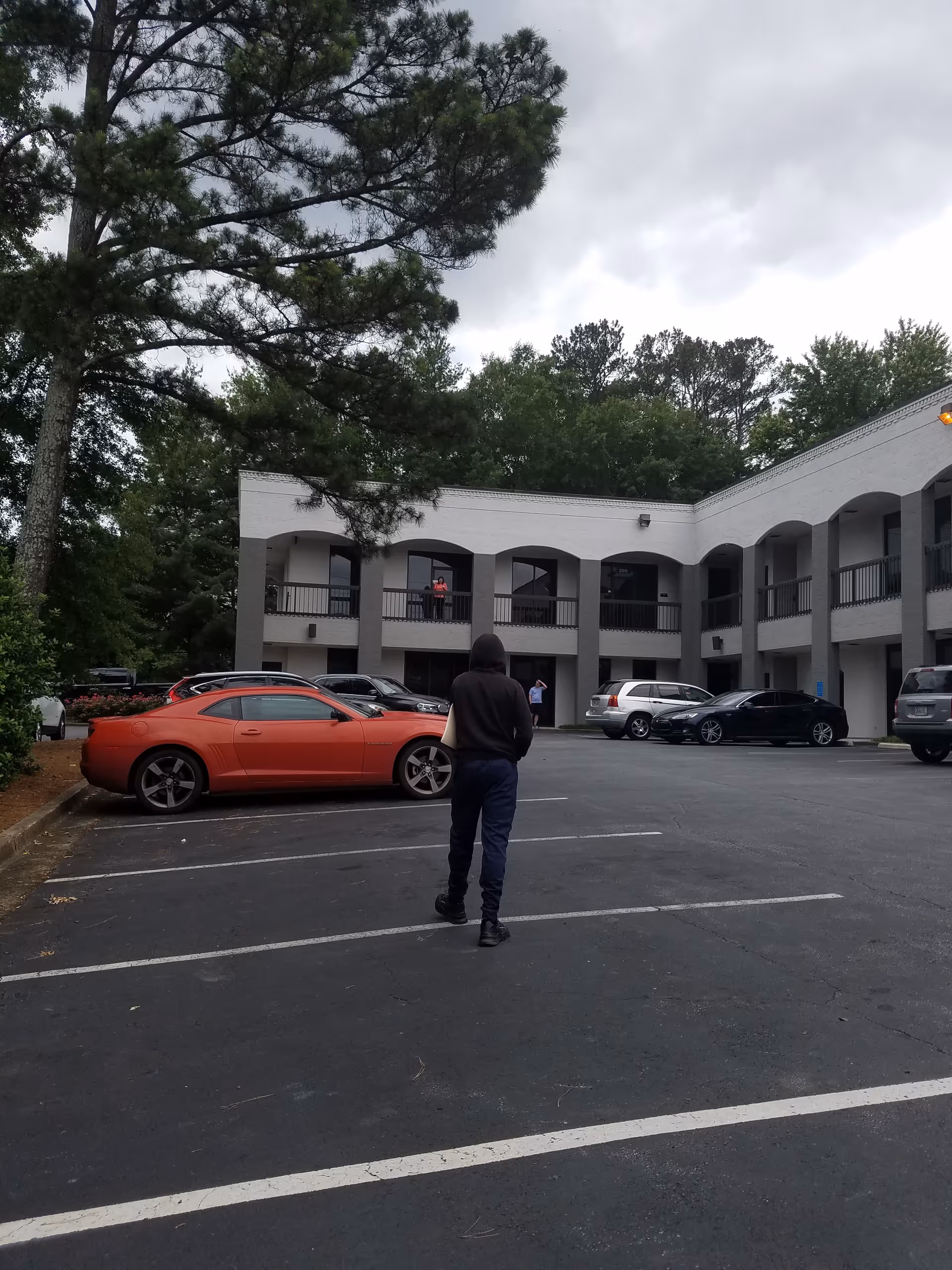 Exterior view of a two-story facility with arches, parked cars, and a person walking.