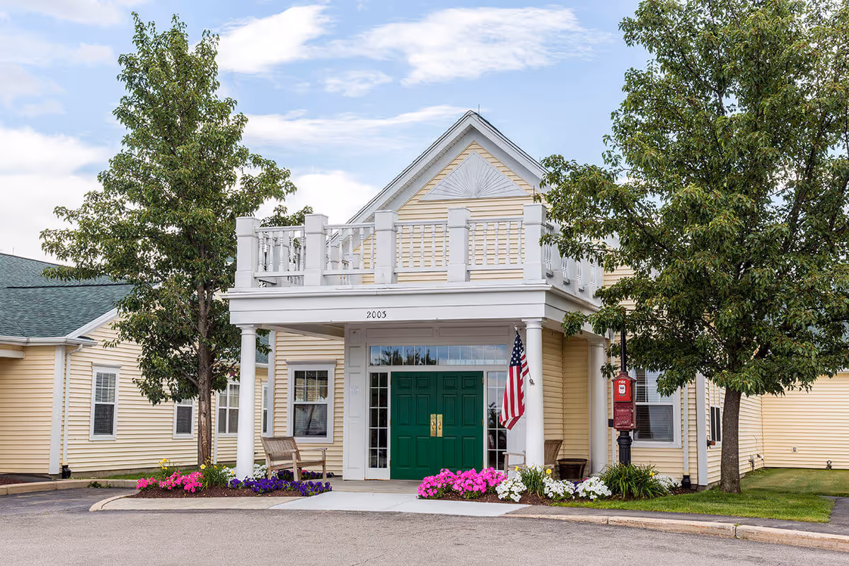 Front entrance of a yellow building with a balcony, green double doors, American flag, and flowerbeds.