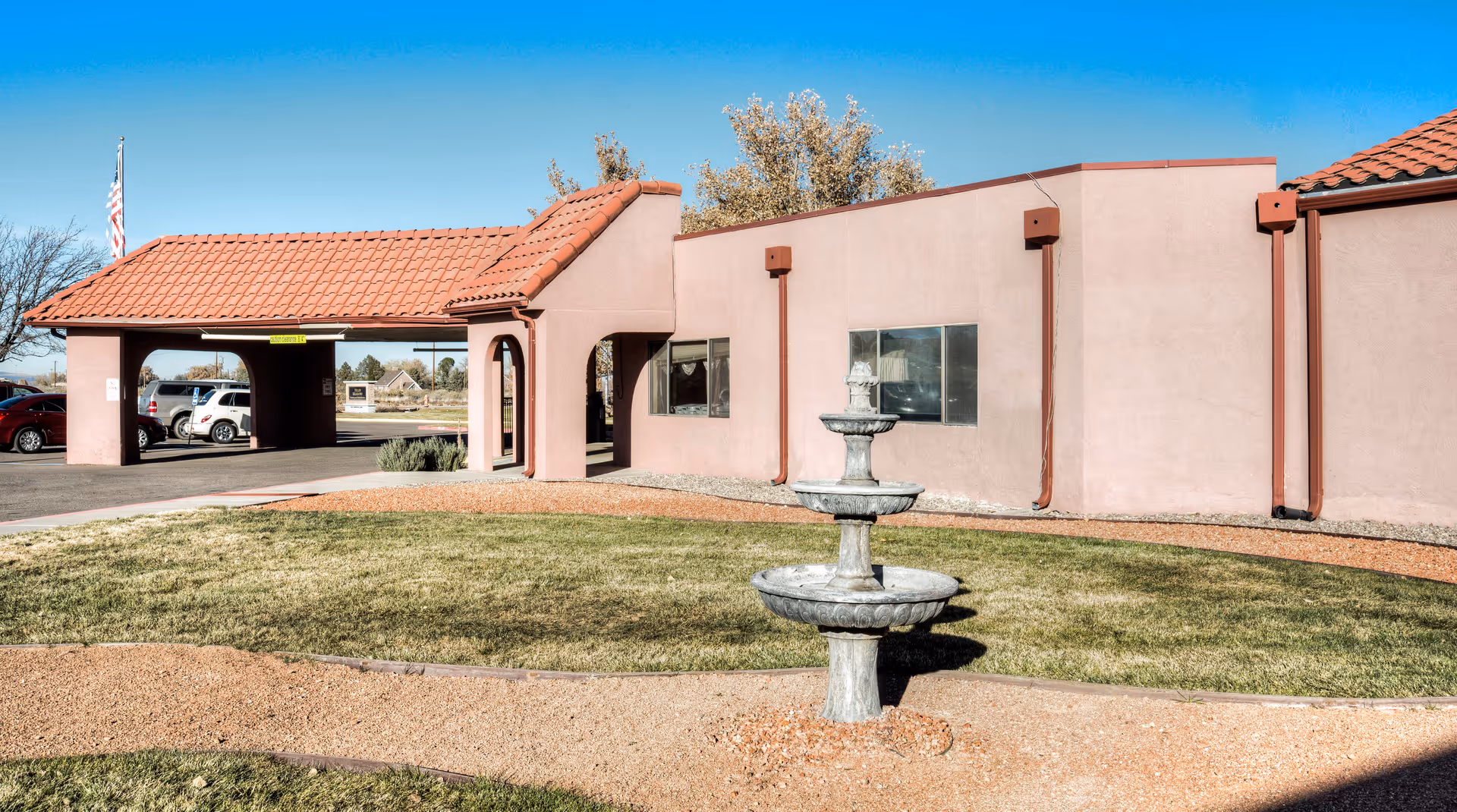 Exterior view of a healthcare and rehabilitation center building with a red tile roof, beige walls, a small fountain in the foreground, a grassy area, and a parking area with several cars under a clear blue sky.