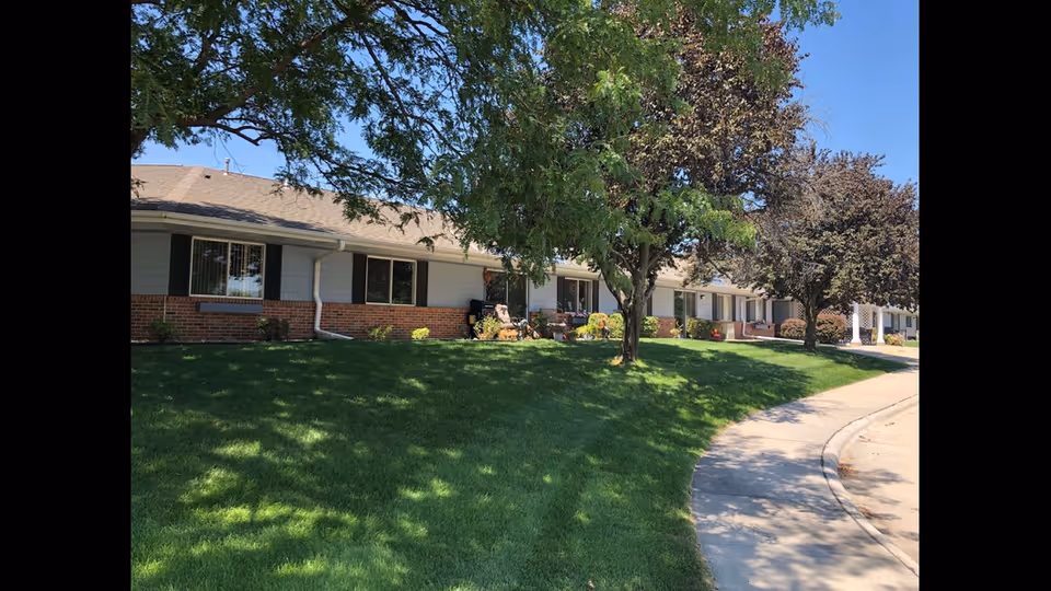 Single-story brick senior living facility with green lawn, trees, and a curved sidewalk.