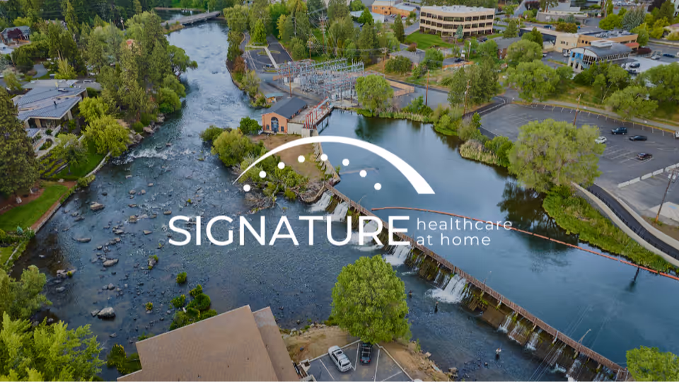 Aerial view of riverfront campus with trees, buildings, and Signature Healthcare at Home logo.