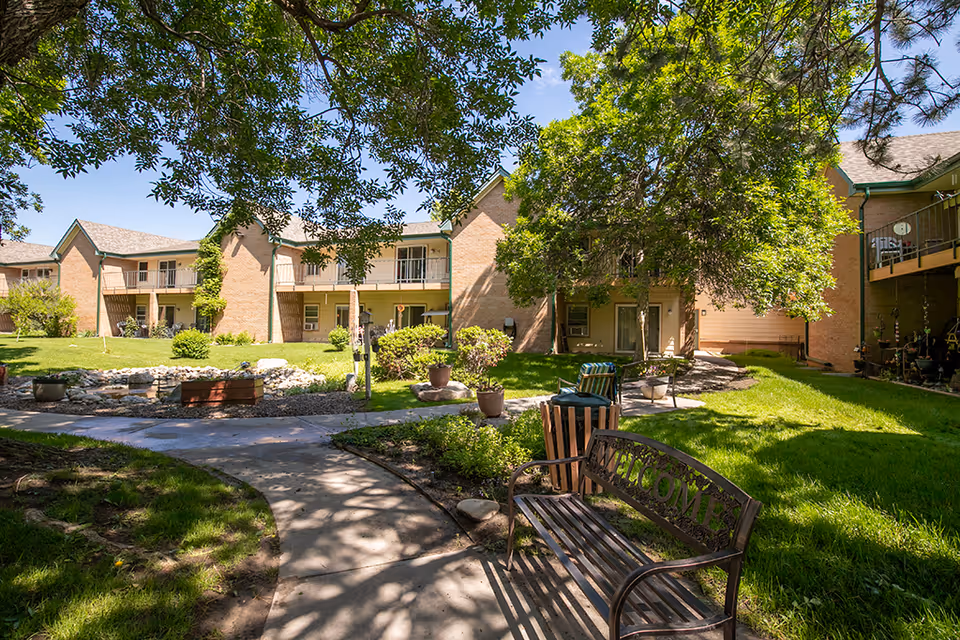 Outdoor courtyard area at Inglenook At Brighton featuring a paved walkway, green grass, trees providing shade, a metal bench with the word 'WELCOME' on the backrest, potted plants, and a two-story brick building with balconies in the background under a clear blue sky.