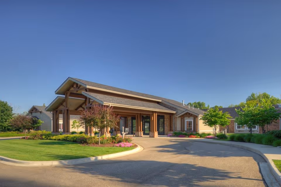 Exterior view of a single-story building with a covered entrance, surrounded by well-maintained landscaping including green grass, small trees, and flower beds under a clear blue sky.