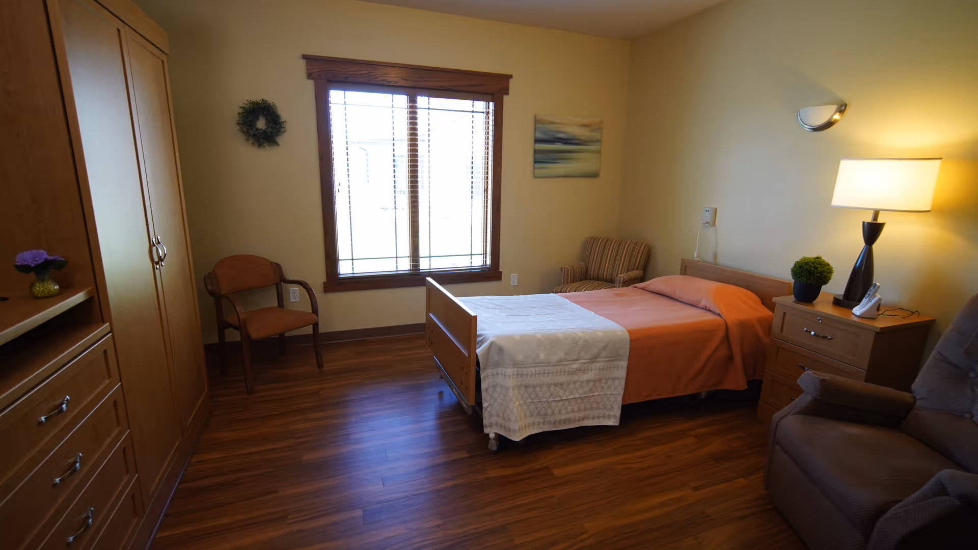 A simple, clean bedroom in a senior living facility with a single bed covered in an orange blanket and white sheet, a wooden nightstand with a lamp and a small plant, a comfortable armchair, a wooden wardrobe, a chair near the window, and a wall decoration. The room has wooden flooring and a large window with blinds letting in natural light.