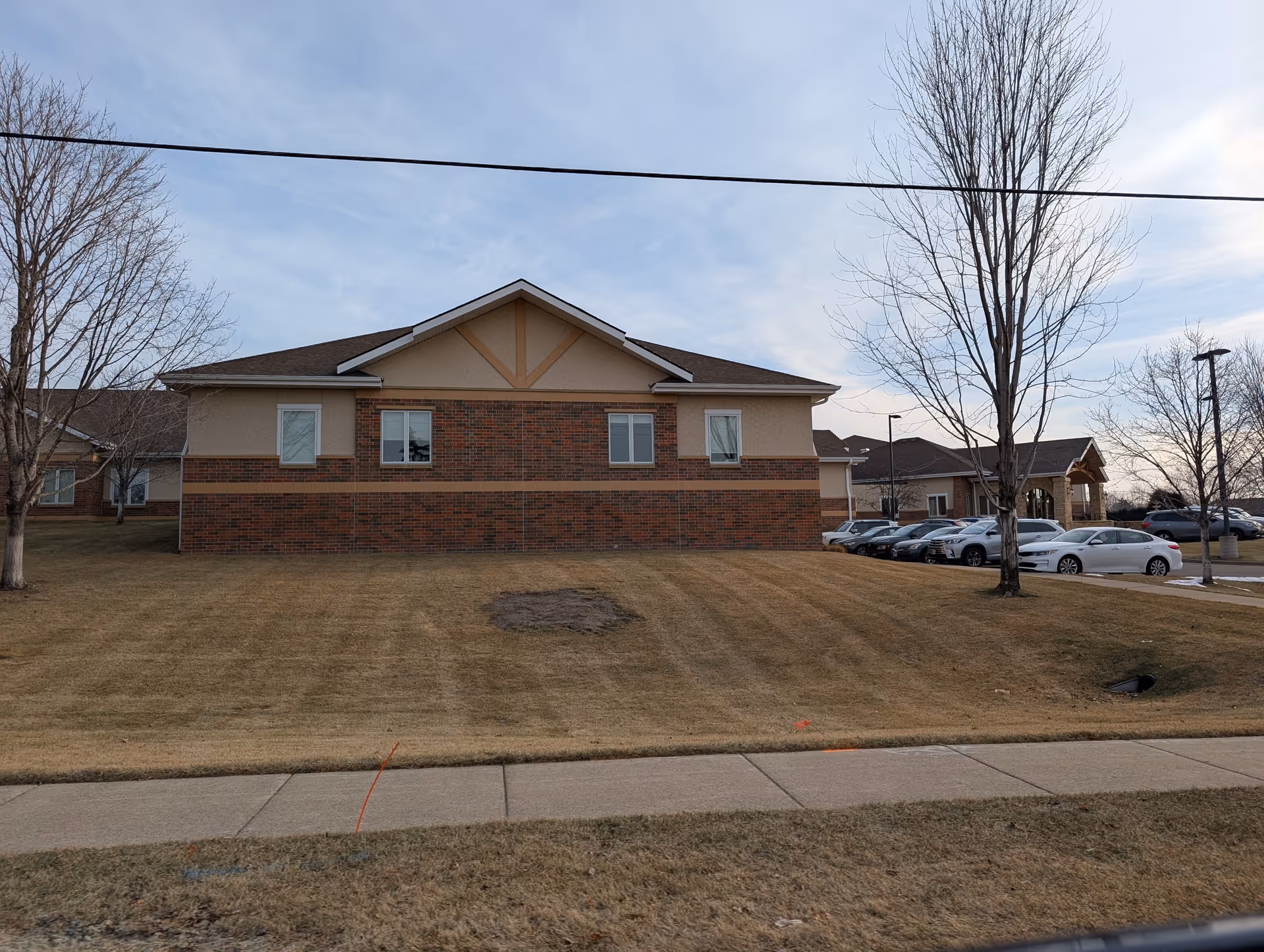 Front exterior of a one-story brick-and-siding senior living building with a grassy lawn, leafless trees, parked cars, and a sidewalk under a cloudy sky.