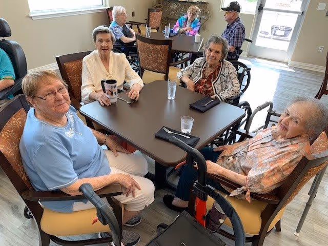 A group of elderly individuals sitting around a table in a communal dining or activity area. They are seated on chairs and some have walkers nearby. The room has large windows letting in natural light and a door leading outside is visible in the background.