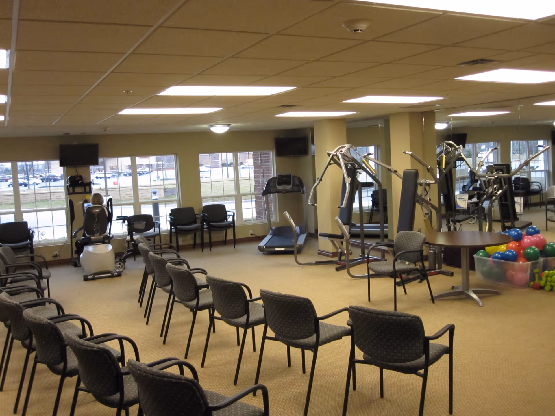 A fitness room with exercise equipment including a treadmill, a recumbent bike, and a multi-station weight machine. Several chairs are arranged in rows facing the equipment. Large windows let in natural light, and a wall mirror reflects the room. A container of colorful exercise balls is visible near a round table.