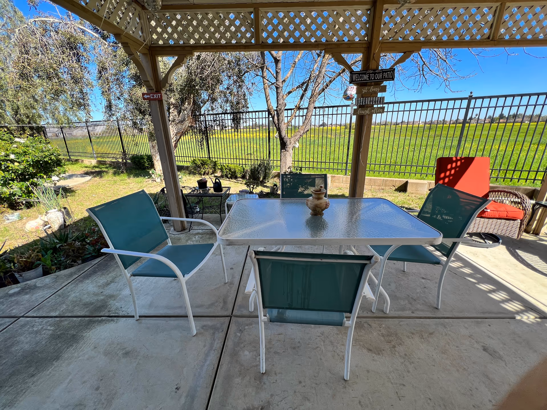 An outdoor patio features a glass table, teal chairs, lattice pergola, and open field beyond.