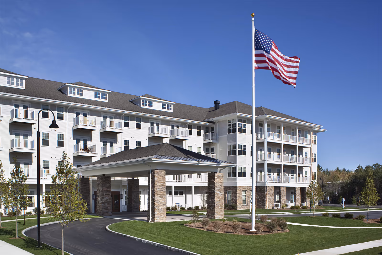 Exterior view of a multi-story senior living facility with white siding and stone accents. The building features multiple balconies and a covered entrance supported by stone pillars. An American flag is flying on a flagpole in front of the building, with a clear blue sky in the background.