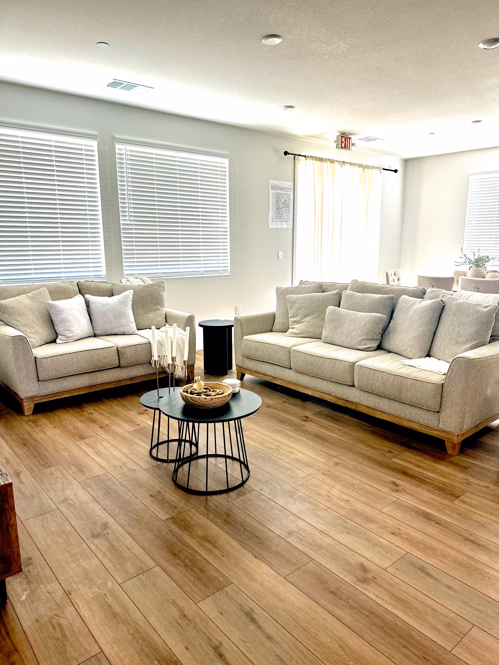 Bright communal living room with beige sofas, wooden floors, and round black coffee tables.