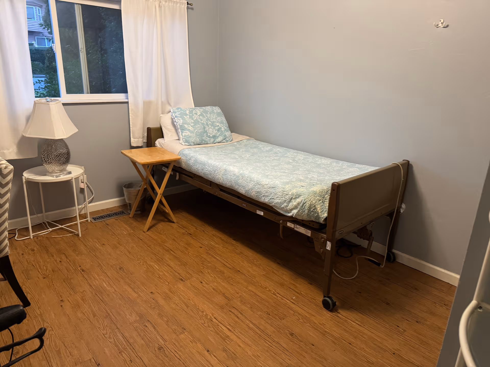 A small bedroom with a hospital-style bed, nightstand, lamp, and window.