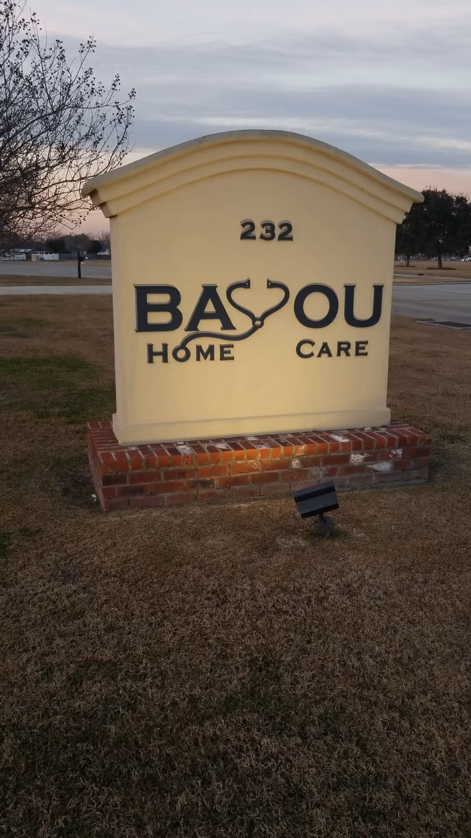 A large curved beige Bayou Home Care sign on a brick pedestal outdoors.