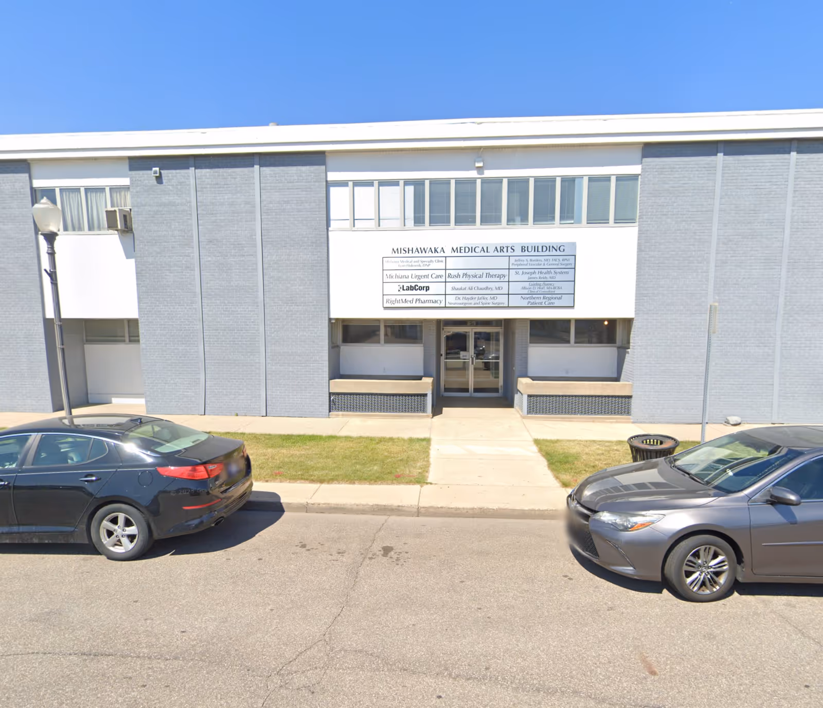 Front exterior of a grey brick building with entrance and parked cars.