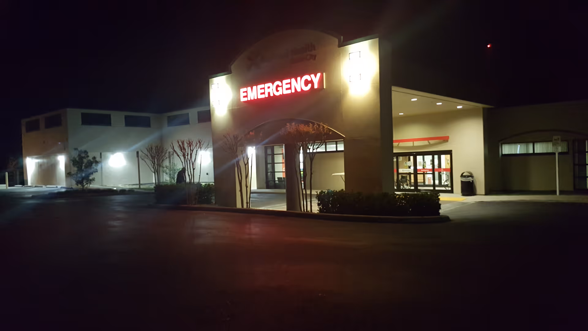 Nighttime exterior of a hospital entrance with a bright EMERGENCY sign.