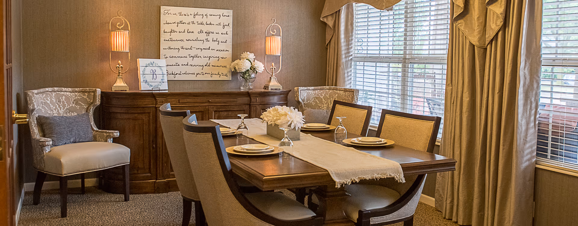 A cozy dining room with a wooden table set with plates and glasses, upholstered chairs, a sideboard with lamps and decor, and a large window with drapes.