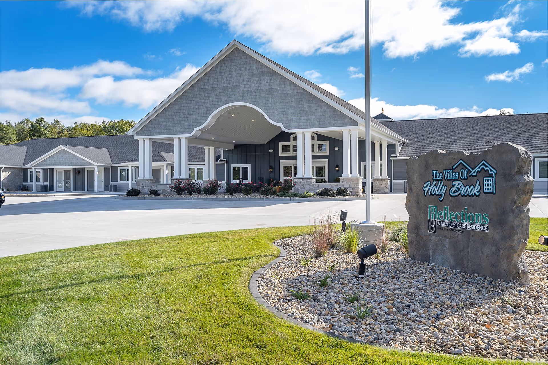 Front entrance of the Villas of Holly Brook assisted living and memory care facility with a covered porte-cochere, landscaped lawn, and a stone sign near the driveway.