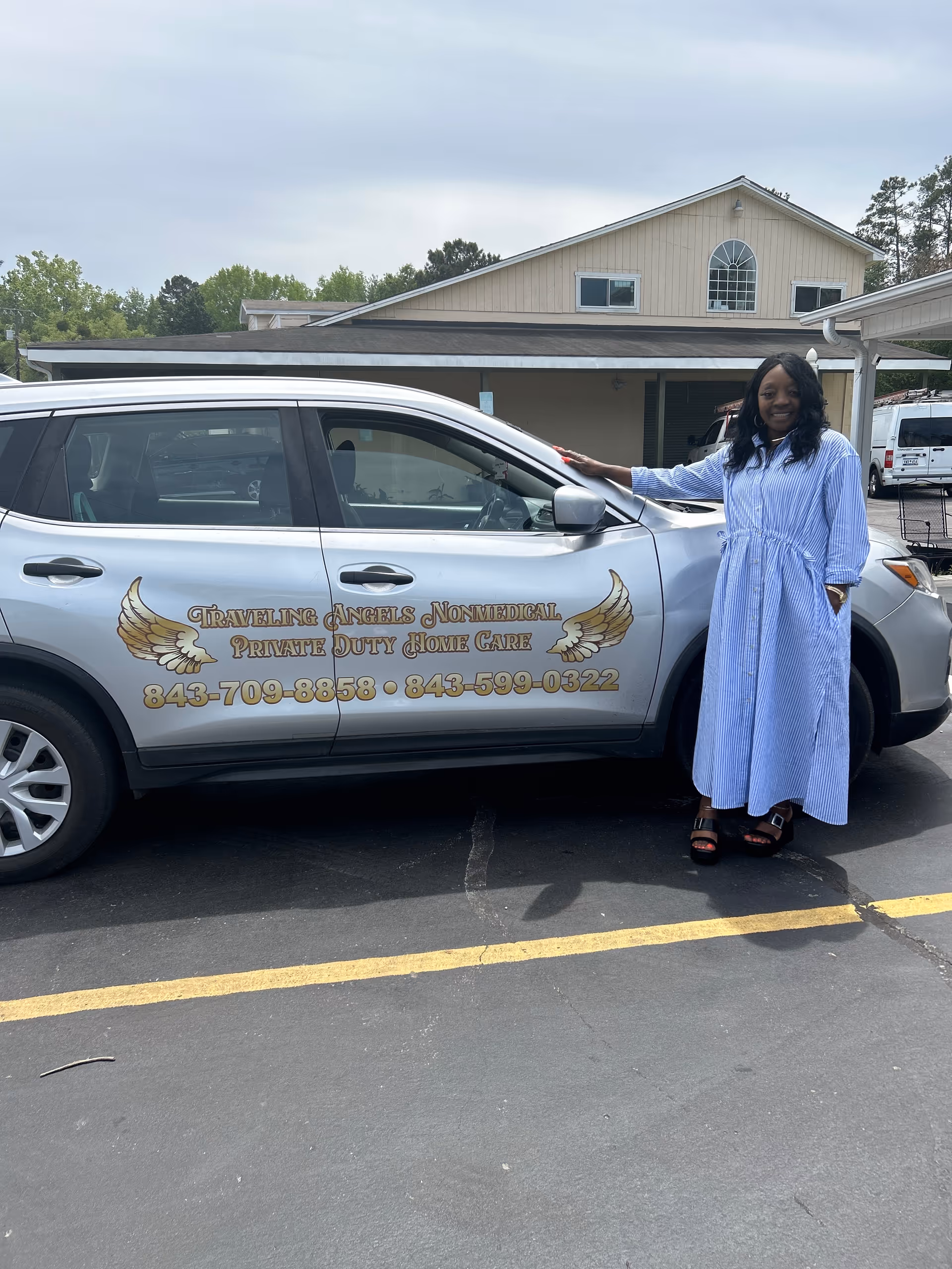 Woman in blue striped dress stands beside a Traveling Angels vehicle outside a facility.