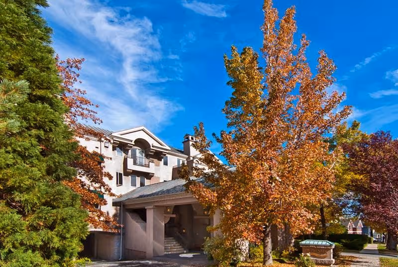 Front entrance of a multi-story senior living building surrounded by trees with autumn foliage under a blue sky.