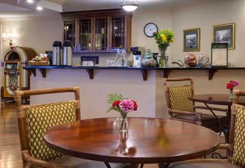 A cozy dining area with round wooden tables and cushioned chairs featuring a patterned fabric. Each table has a small vase with fresh flowers. In the background, there is a counter with coffee dispensers, glassware, and decorative items, along with a wall clock and framed pictures on the wall.