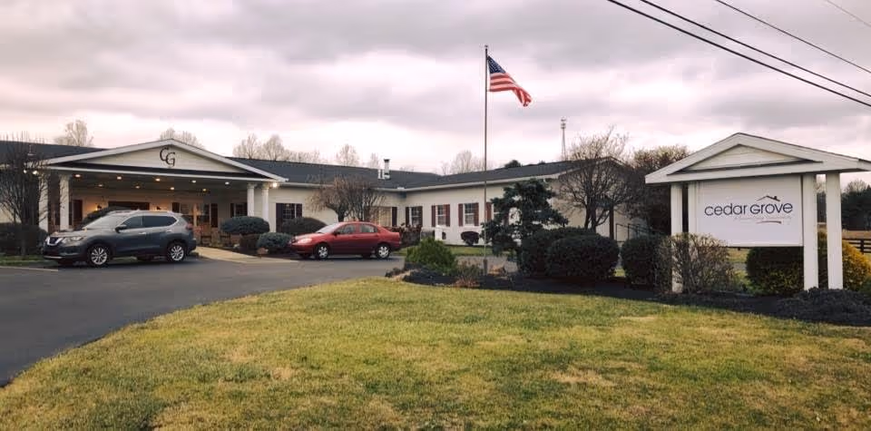 Exterior view of Cedar Grove facility showing a single-story building with a covered entrance, two parked cars, an American flag on a flagpole, and a sign with the facility name on a grassy lawn under a cloudy sky.