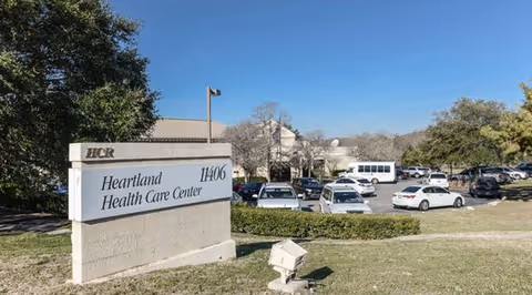 A sign reading 'Heartland Health Care Center 11406' in front of a parking lot with cars and the facility building under a clear blue sky.