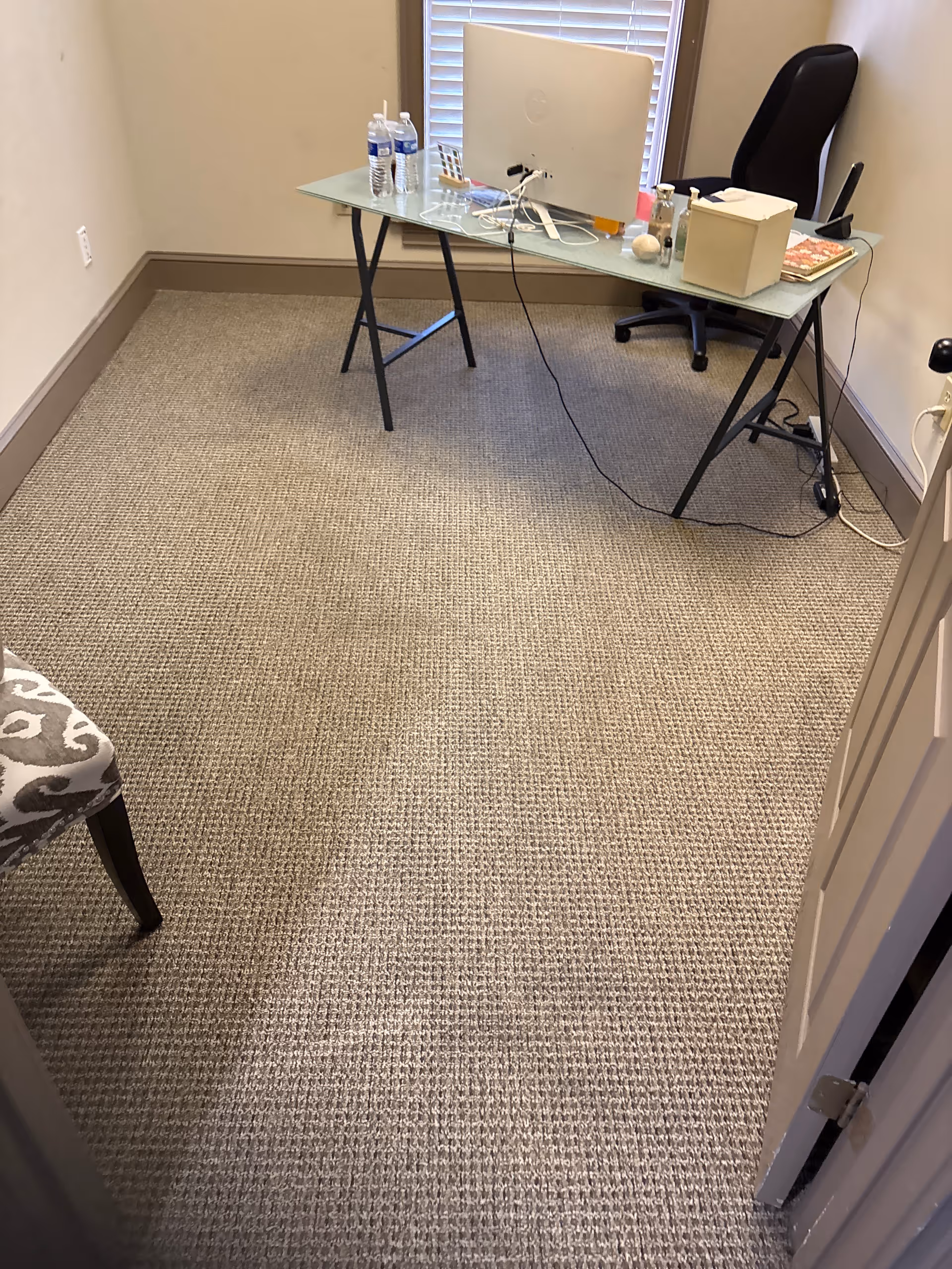 Small glass-topped desk with computer, chair, and supplies in a carpeted room.