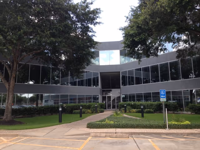 A modern glass-front facility entrance surrounded by trees and landscaped lawn.