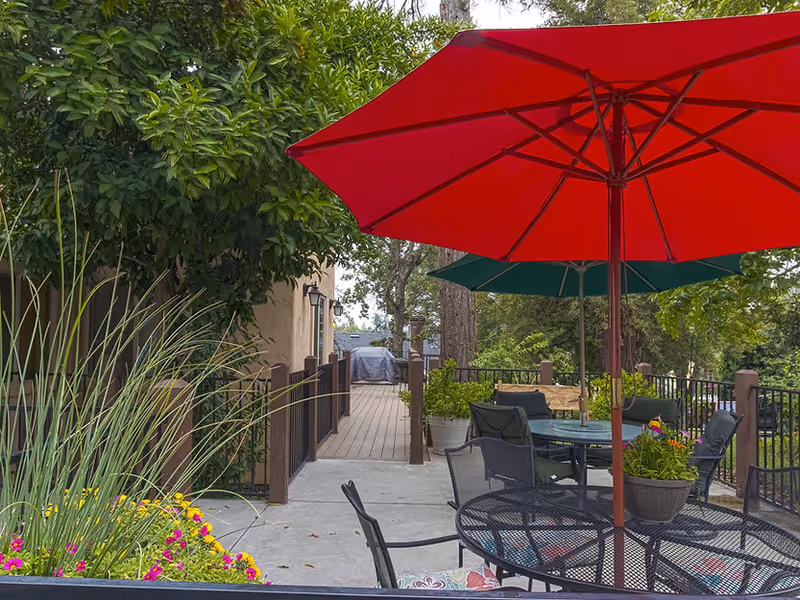 Patio at Chateau Senior Living with metal tables and chairs under red and green umbrellas, potted flowers, and a wooden walkway surrounded by trees.