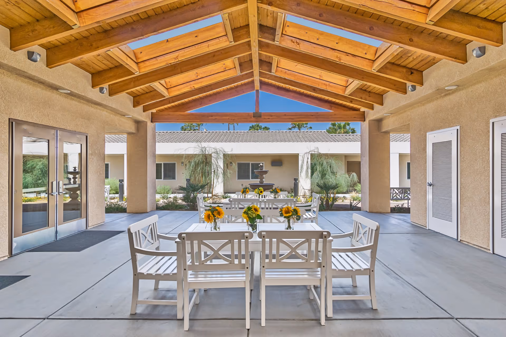 Covered outdoor seating area with white benches and tables adorned with small vases of sunflowers, under a wooden beam ceiling with skylights. In the background, there is a courtyard with plants and a water fountain, surrounded by a beige building with windows and doors.