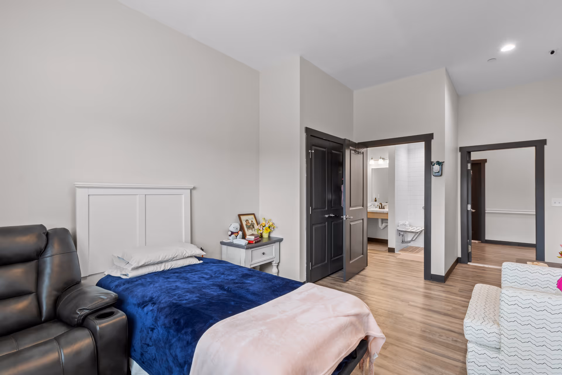 A furnished senior living bedroom with a bed, recliner, nightstand, and doorway to a bathroom.