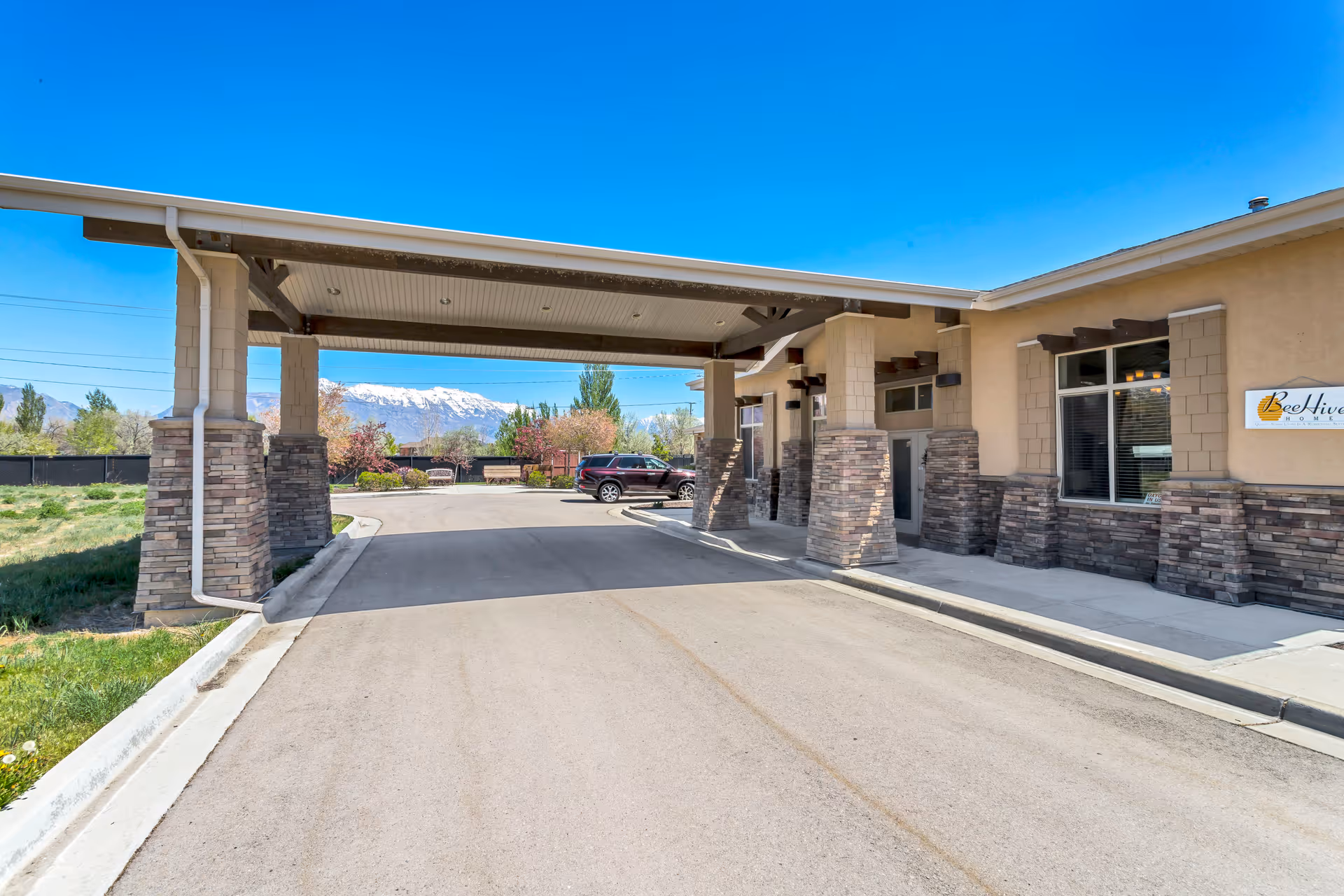 Entrance area of BeeHive Homes of Lehi facility with a covered driveway supported by stone pillars, a parked car, and a clear view of snow-capped mountains in the background under a bright blue sky.