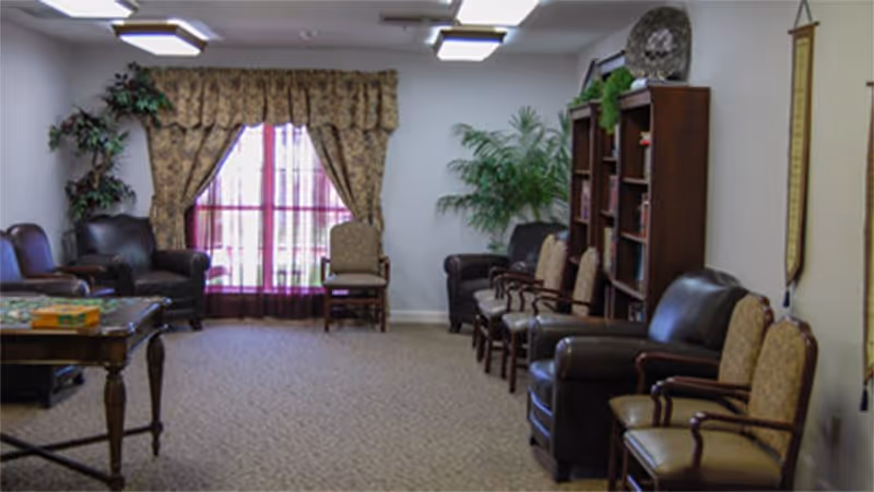 A cozy common area with several upholstered chairs arranged along the walls, a wooden bookshelf filled with books, a table with a board game, and large windows with patterned curtains letting in natural light. There are also two potted plants adding greenery to the room.