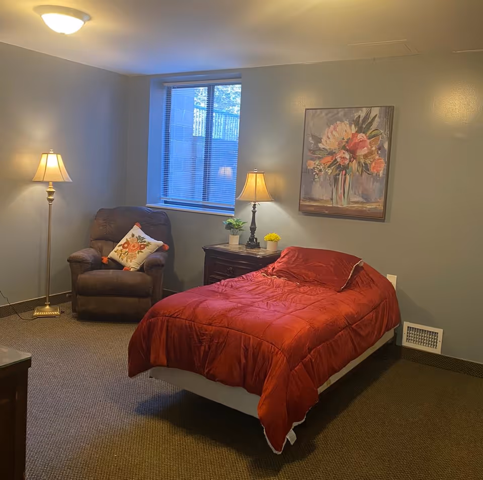 Cozy bedroom with red comforter, armchair, nightstand, lamps, window, and floral wall art.