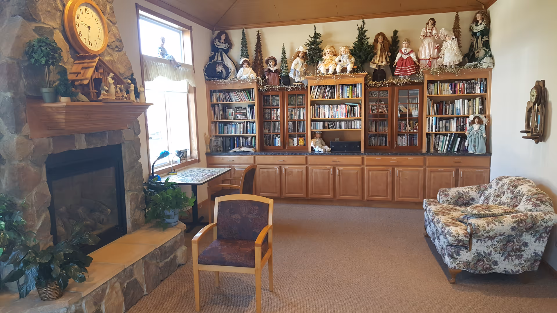 Cozy living room with stone fireplace, tall bookcases, dolls, and floral armchair.