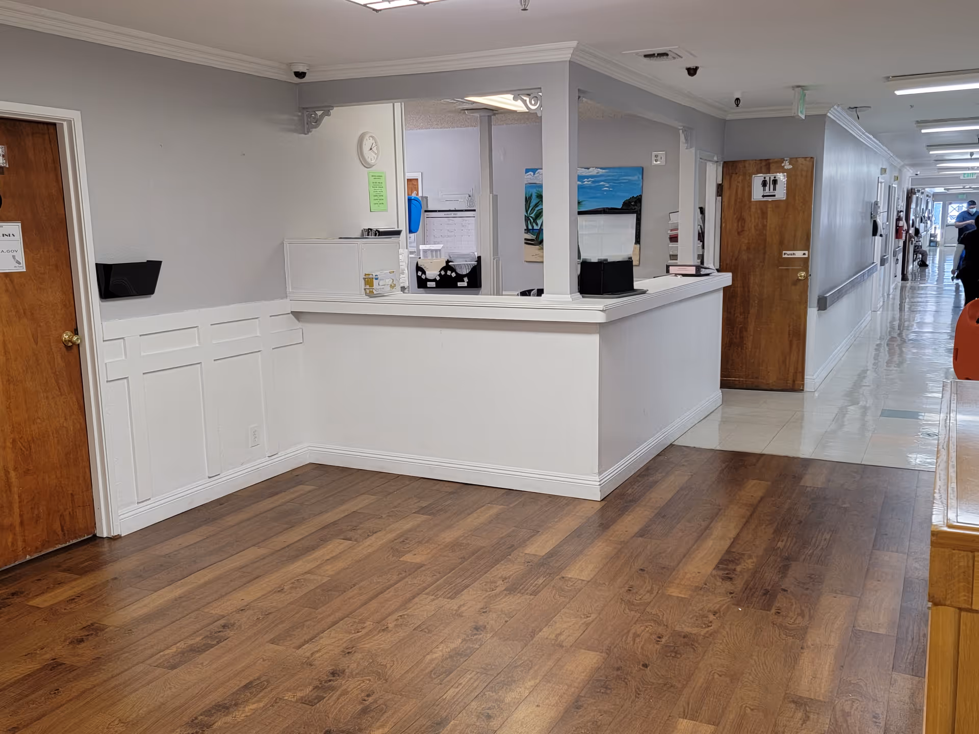 Interior view of a reception or nurse station area in Pasadena Grove Health Center with a wooden floor section in the foreground, a white counter with a small window opening, and a hallway extending to the right with doors and a restroom sign visible.