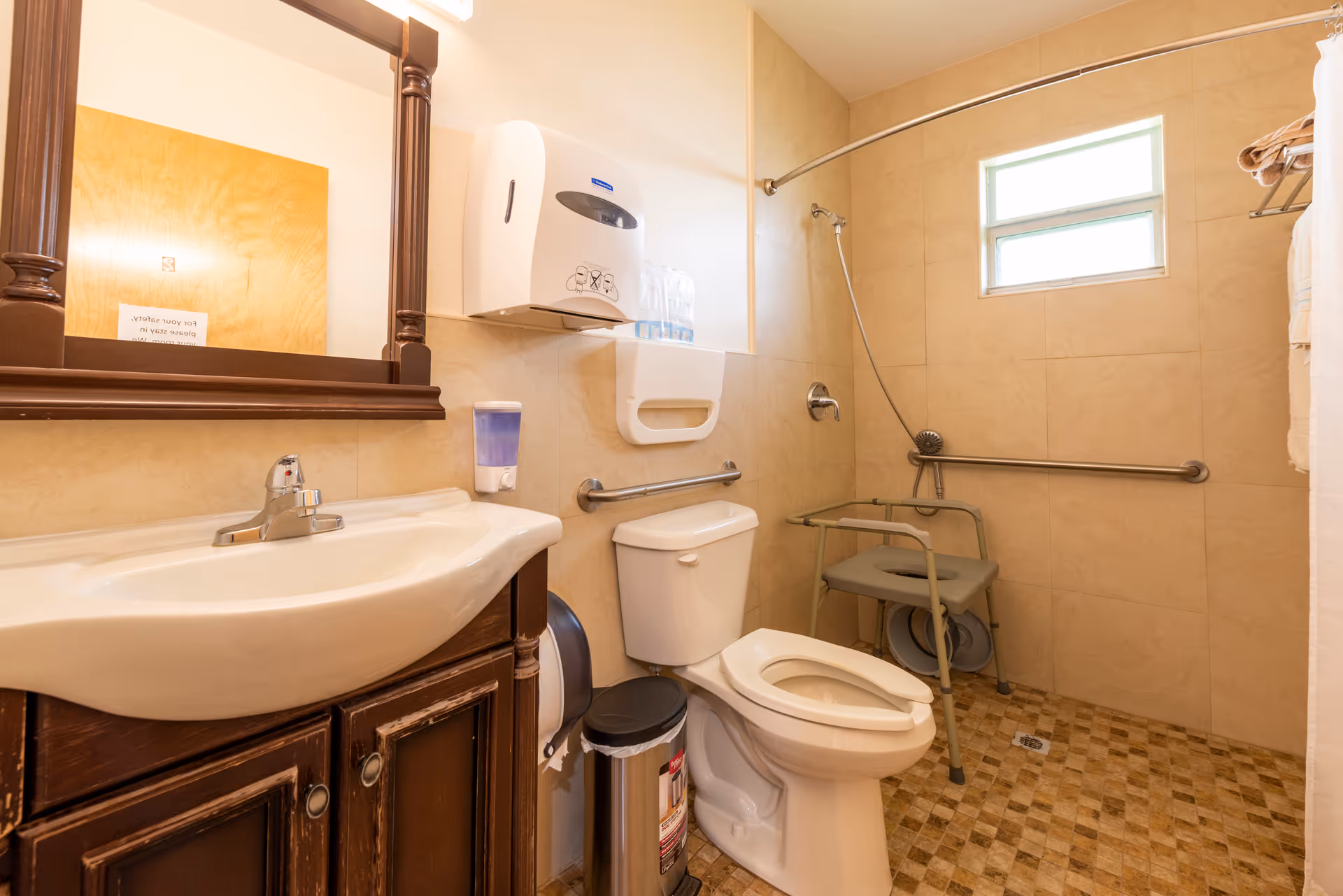 Accessible senior living bathroom with sink, mirror, toilet, grab bars, and a shower chair.
