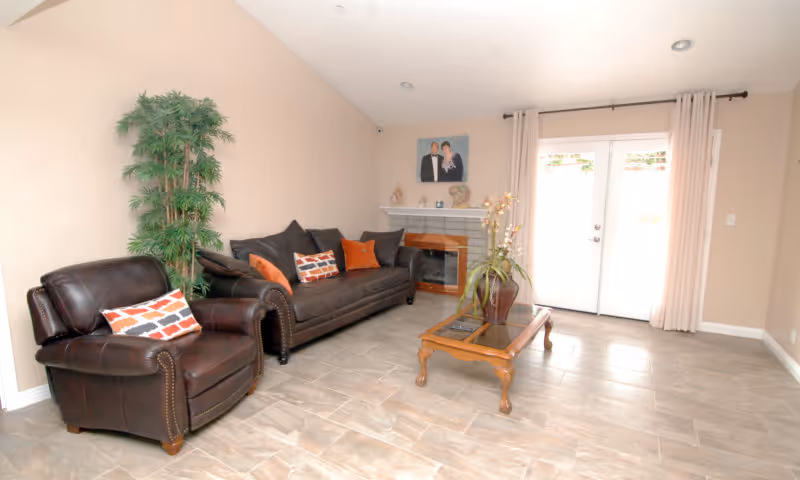 A living room with a dark brown leather armchair and matching sofa adorned with decorative pillows. There is a tall green plant in the corner, a wooden coffee table with a glass top and a vase with flowers, a fireplace with a mantel displaying small decorative items, and a framed photo above it. Double glass doors with light curtains allow natural light into the room.
